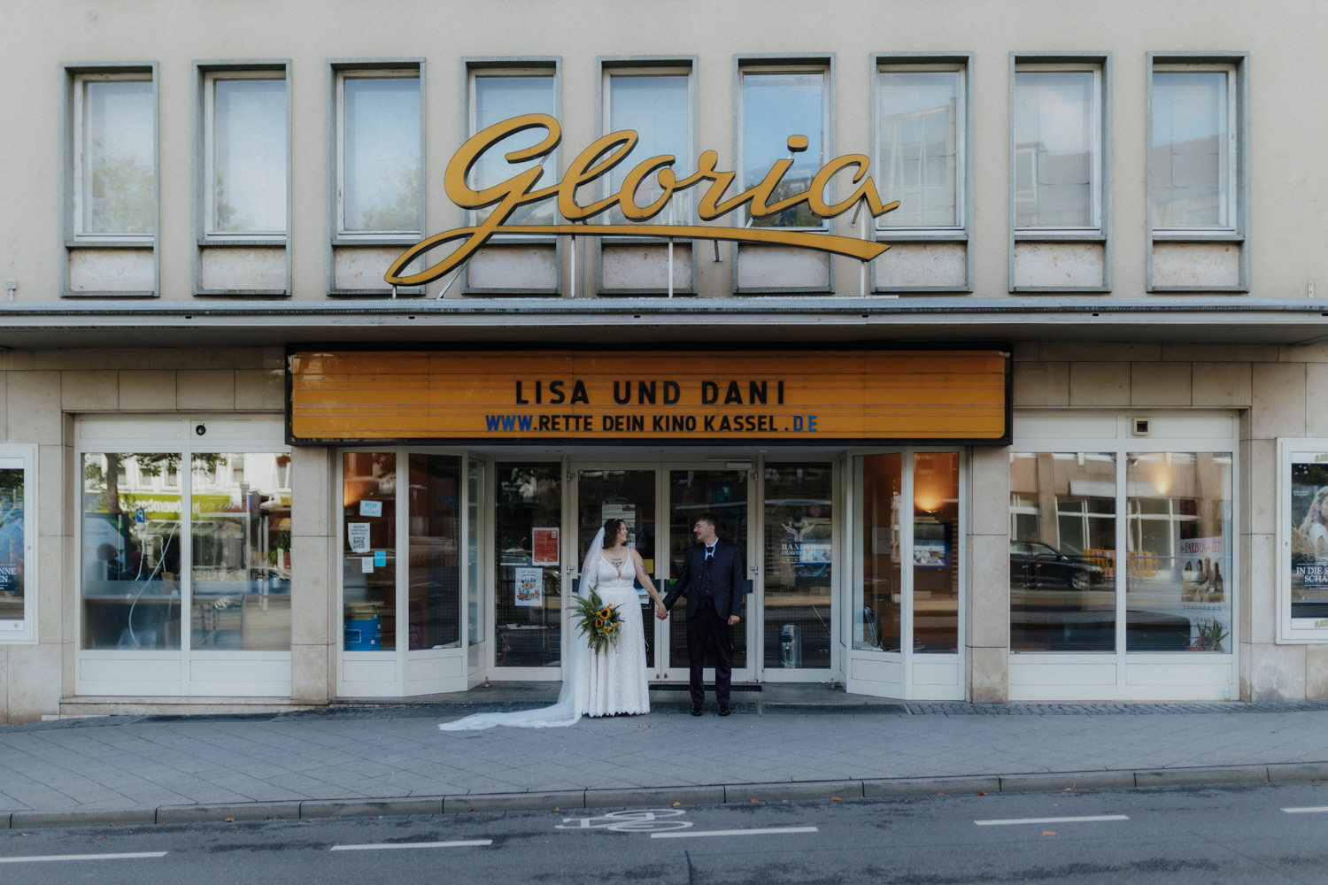 Hochzeit vor Kinoeingang Kassel – Brautpaar hält Hände vor Gloria