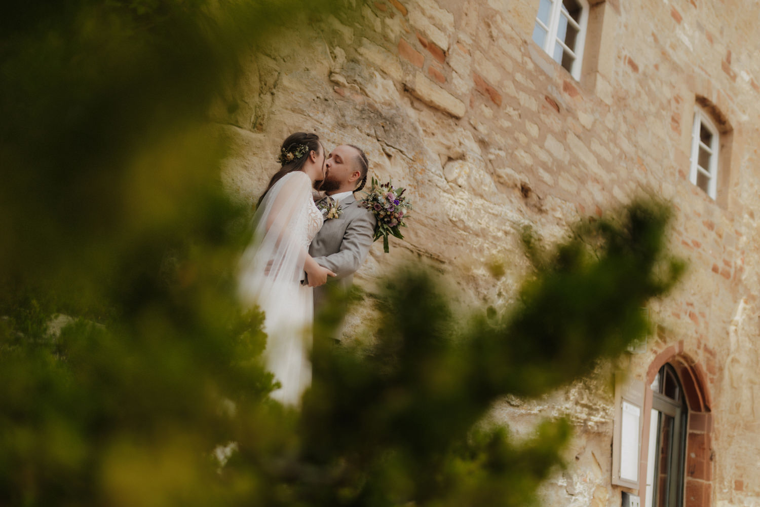 Brautpaar küsst sich vor Schlossmauer Waldeck durch grüne Blätter